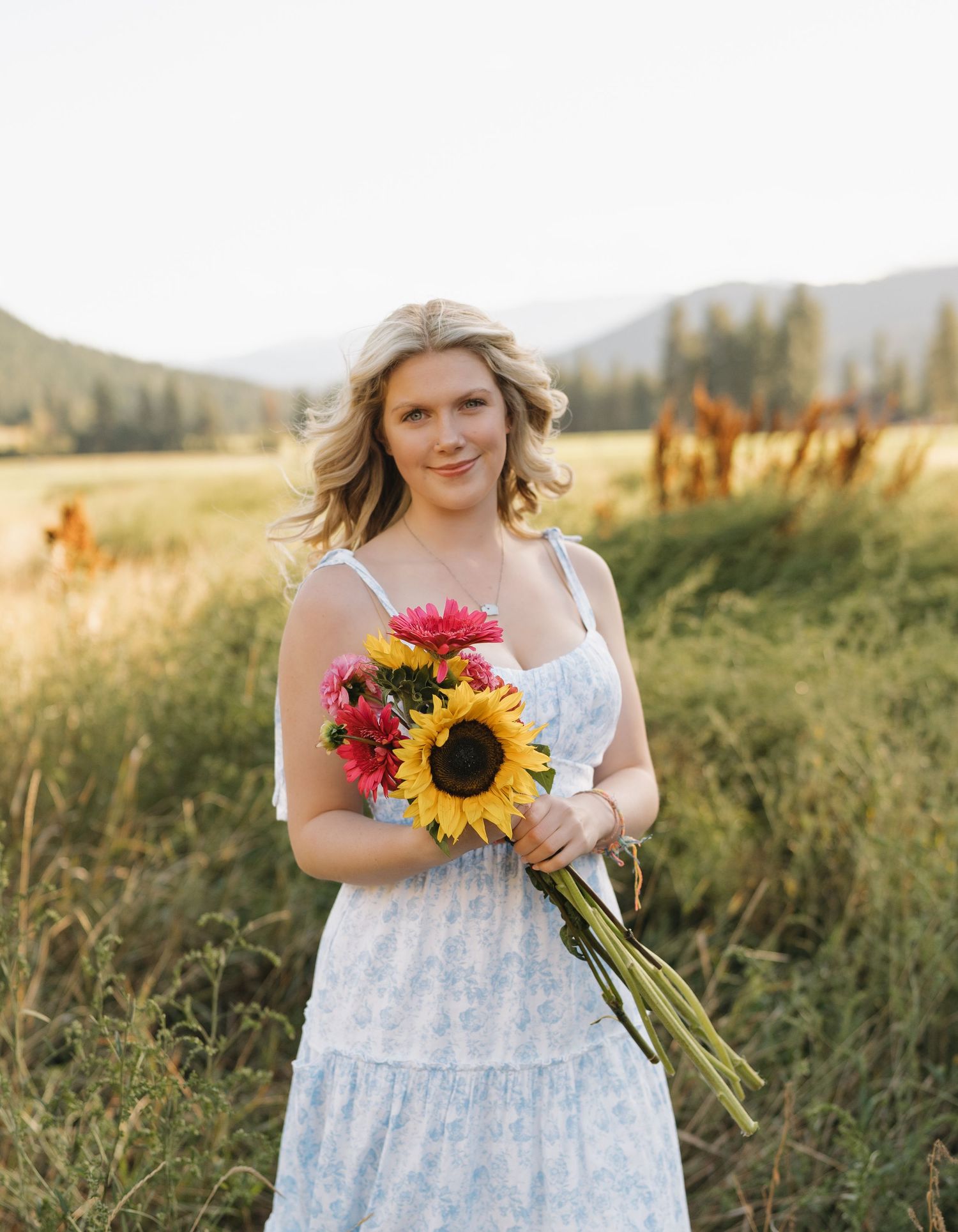 Person in white dress holding sunflower bouquet in sunny mountain field.