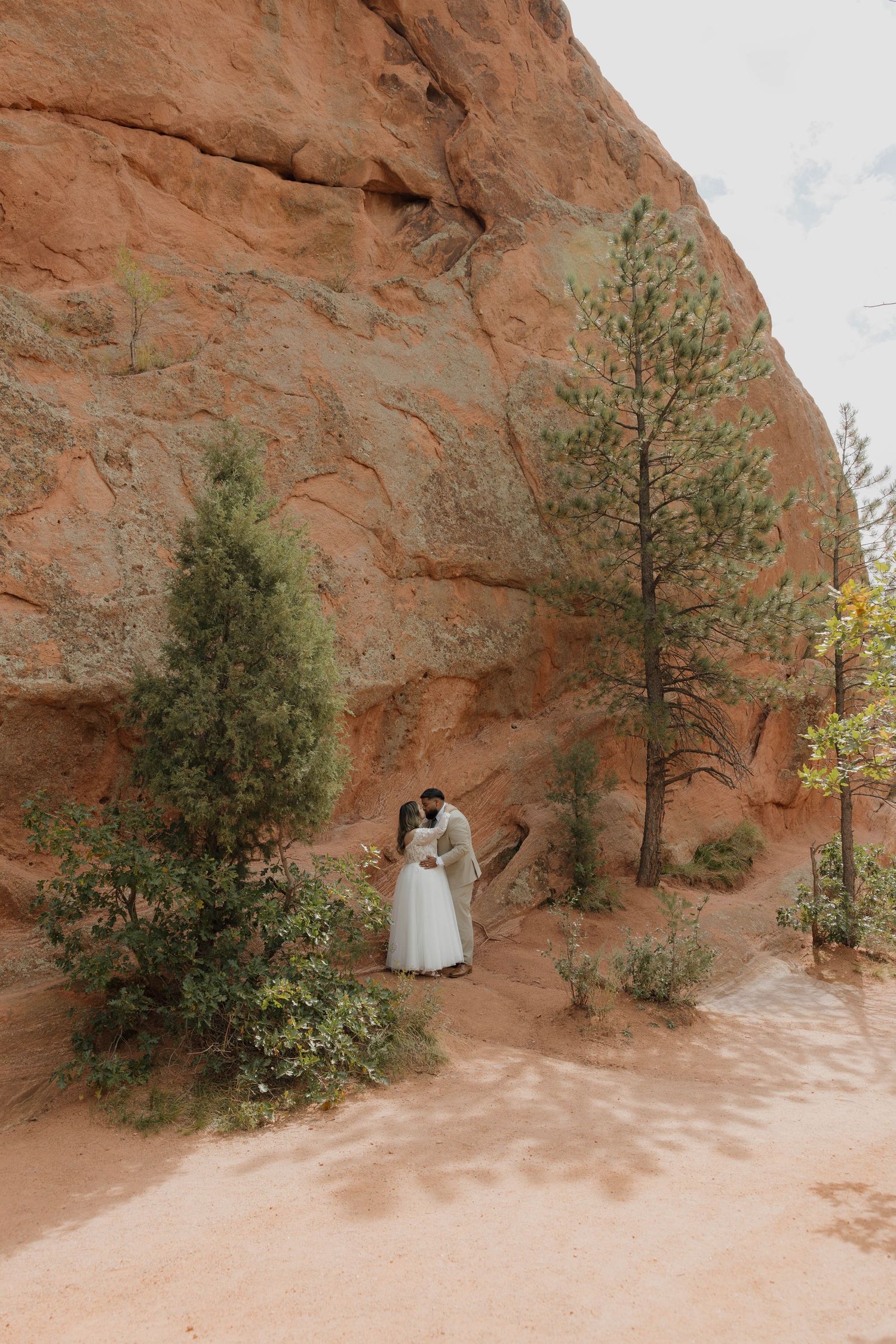 A romantic couple stands against red rock formations in a desert landscape with evergreen trees.