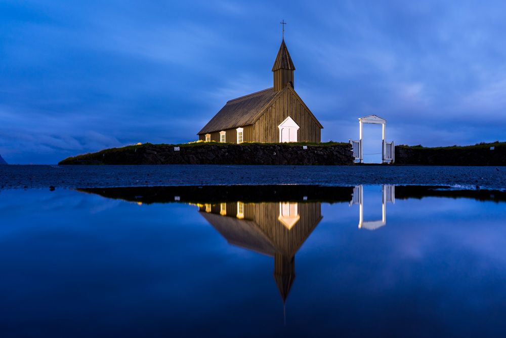 A small wooden church with steeple reflects perfectly in calm waters at twilight against a deep blue sky.
