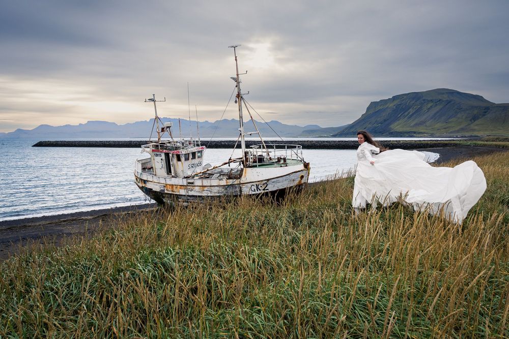 Scenic panoramic view of a fishing boat anchored near a rugged coastline with mountains and dramatic cloudy skies.