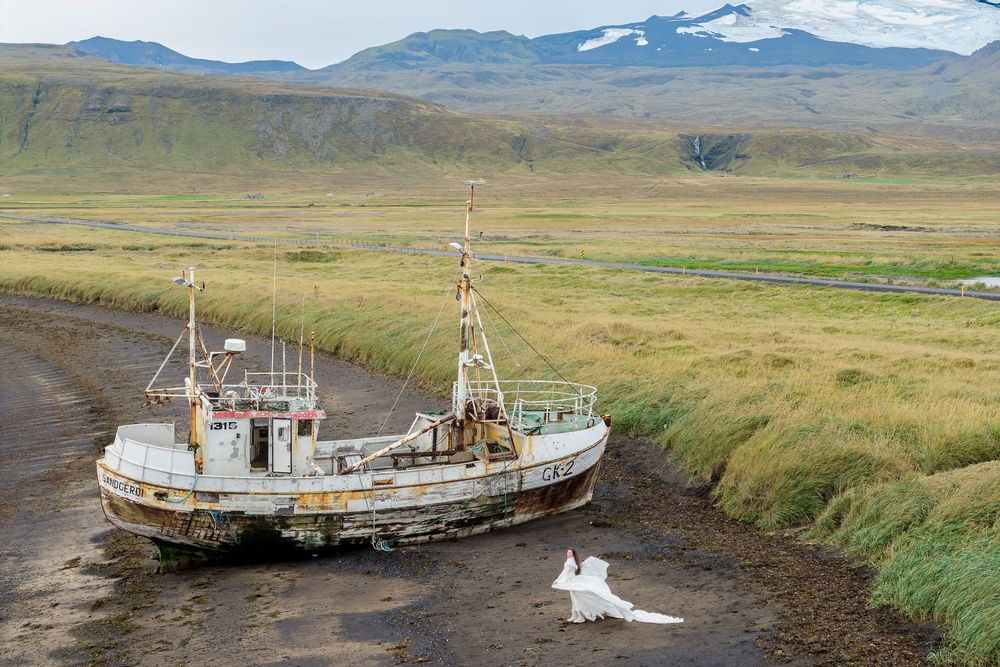 Seven abandoned fishing boats rest on a grassy shoreline with mountains in the distance under cloudy skies.