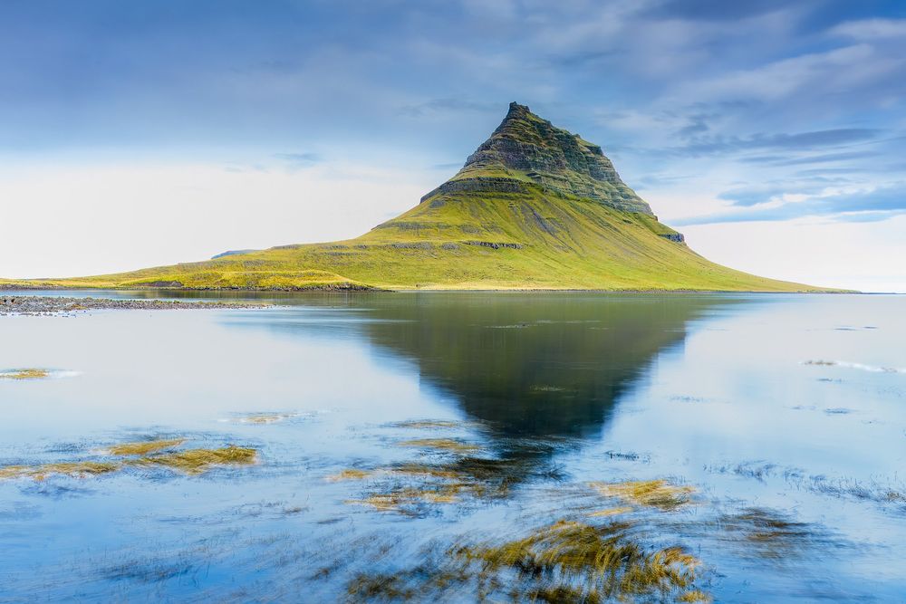 Panoramic view of a majestic mountain peak reflecting in calm waters under blue skies in Iceland's rugged landscape.