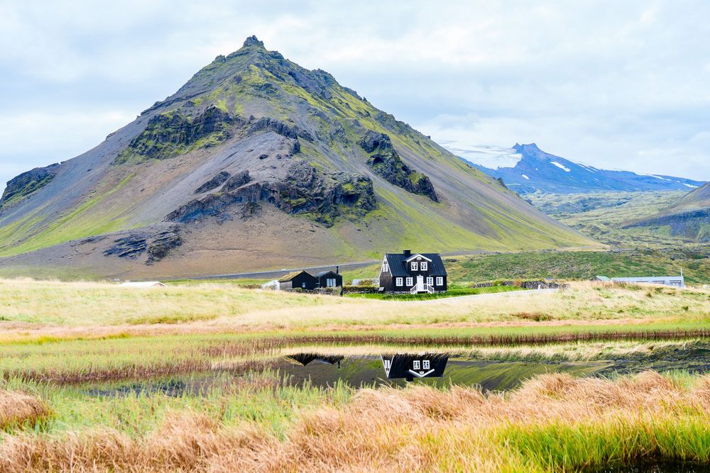 A panoramic view of rugged mountain peaks and green valleys in the Icelandic countryside with scattered rural houses.