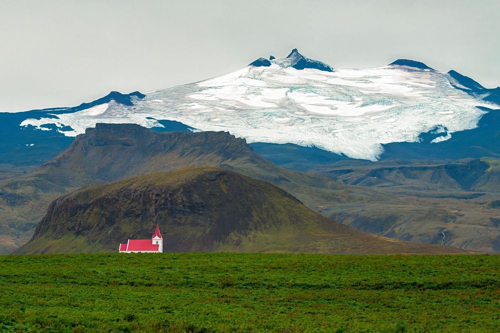Snow-capped mountains tower over rugged Icelandic terrain with a solitary red-roofed building in the distance.