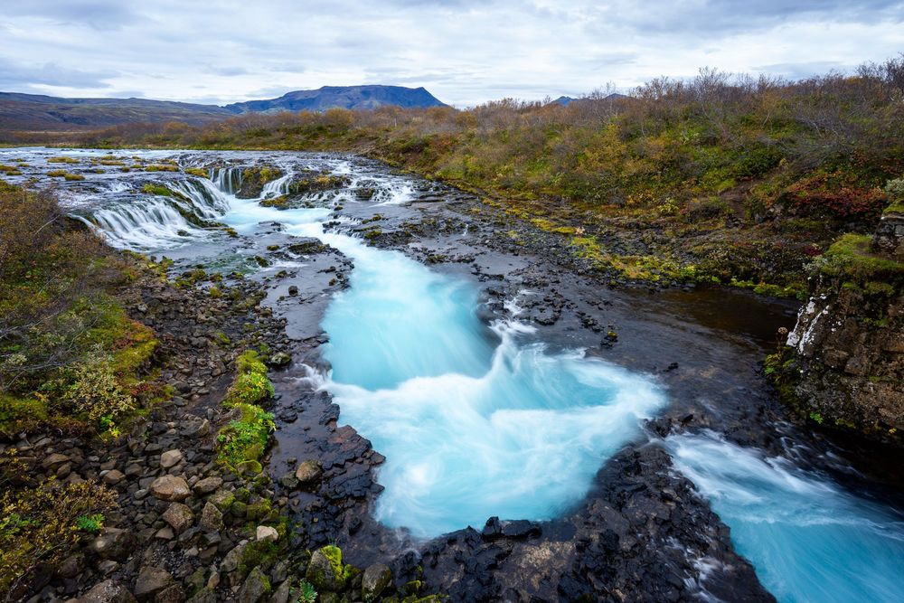 Turquoise glacial water cascades over rocky terrain at Bruarfoss waterfall in Iceland's rugged landscape.