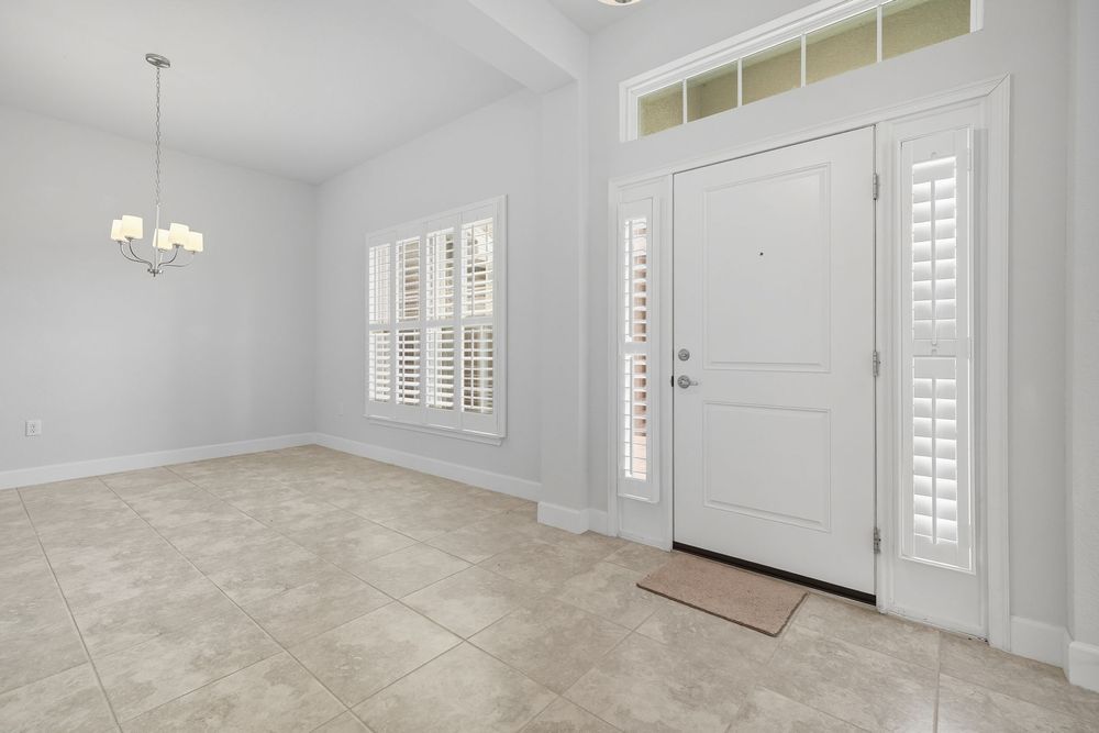 Bright empty room with white walls, plantation shutters, tile flooring and a chandelier near the front entrance.