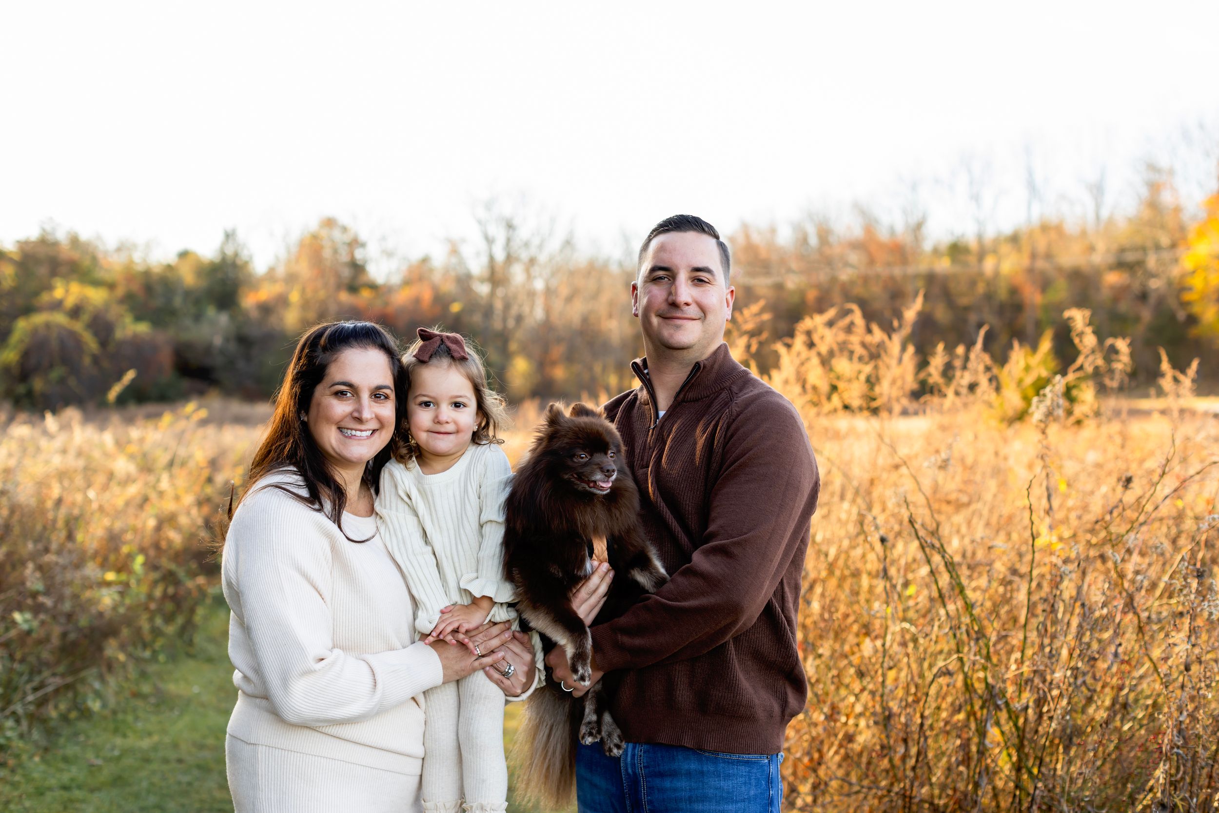 A family and their black dog pose together in a golden autumn field at sunset.