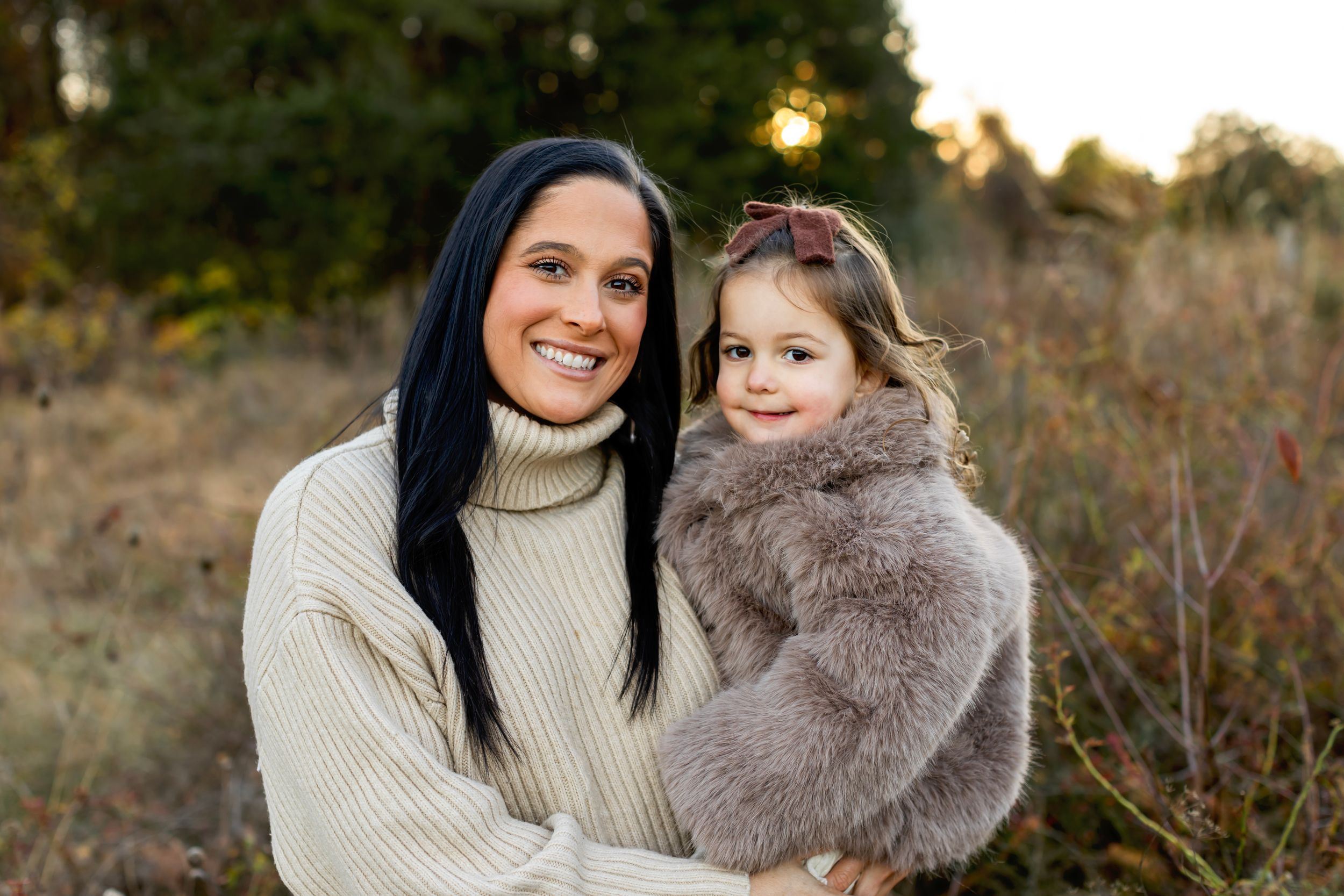 Two people wearing warm sweaters pose together outdoors during golden hour in a natural field setting.