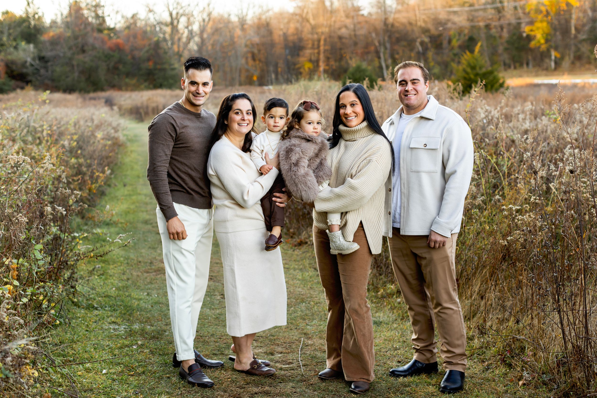 A group of people wearing coordinated brown and white outfits pose together in an autumn field at sunset.