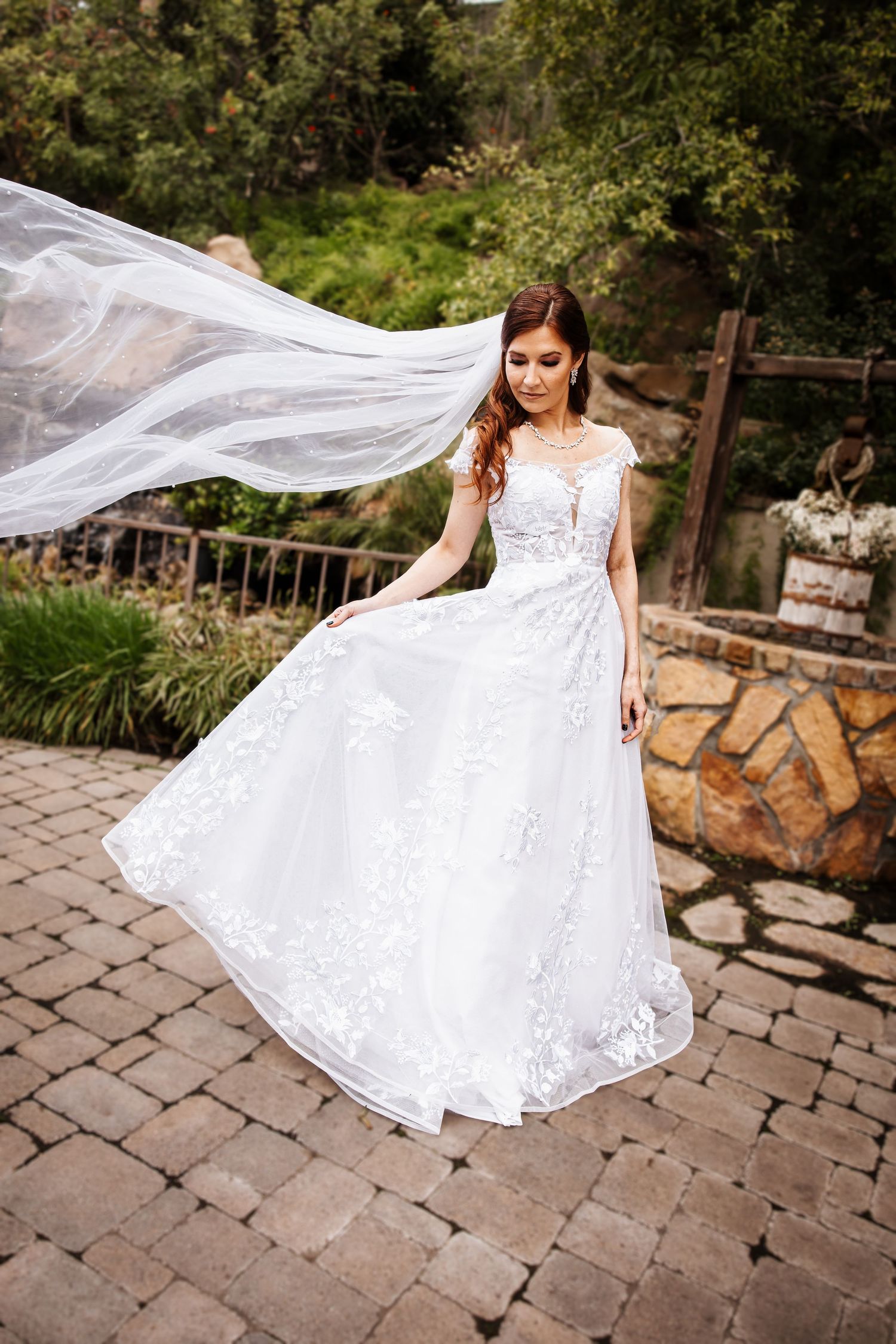 Bride in lace ball gown with veil flowing in wind on stone patio surrounded by greenery.