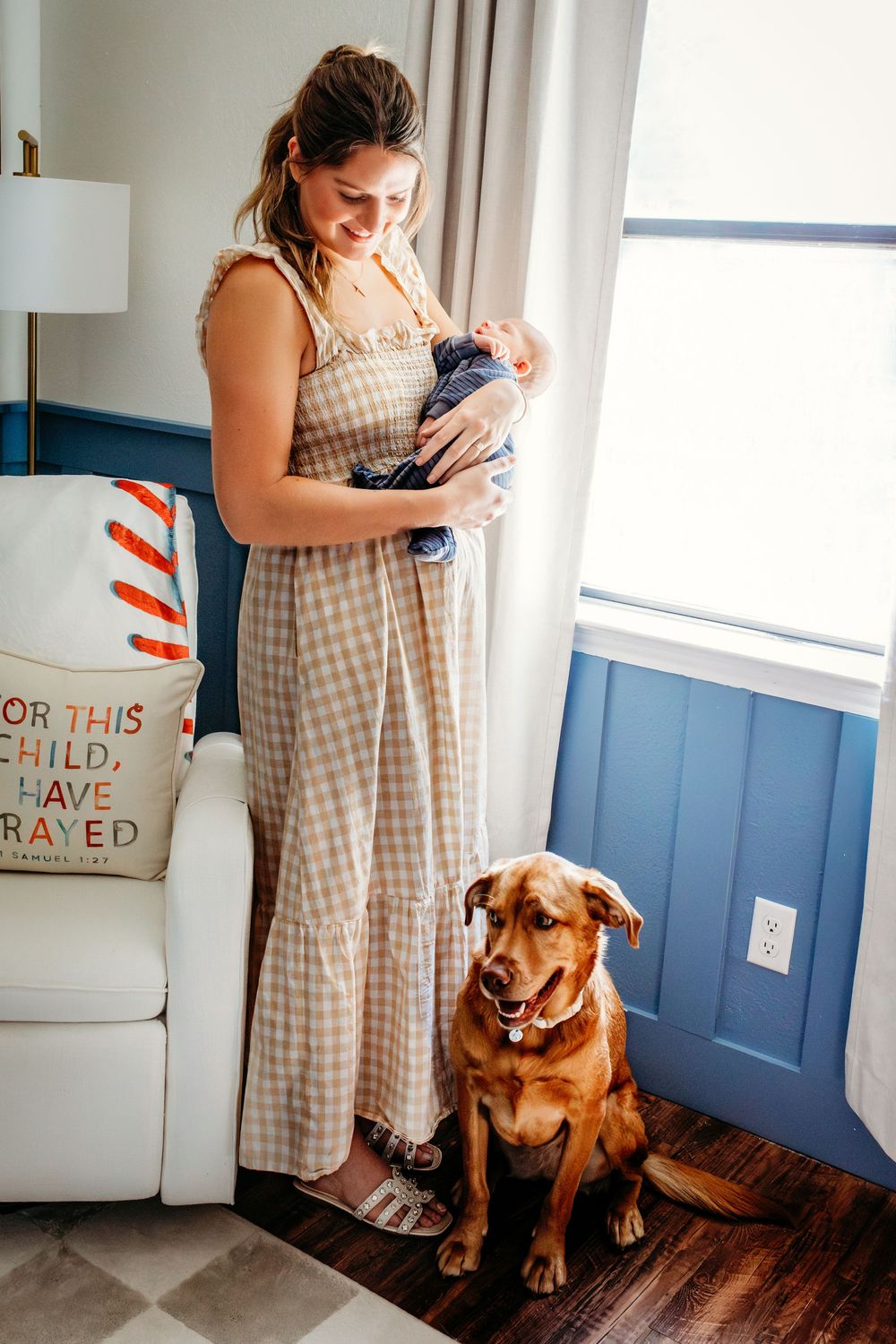 A brown dog sits patiently on hardwood floors next to blue walls while someone holds a newborn baby.