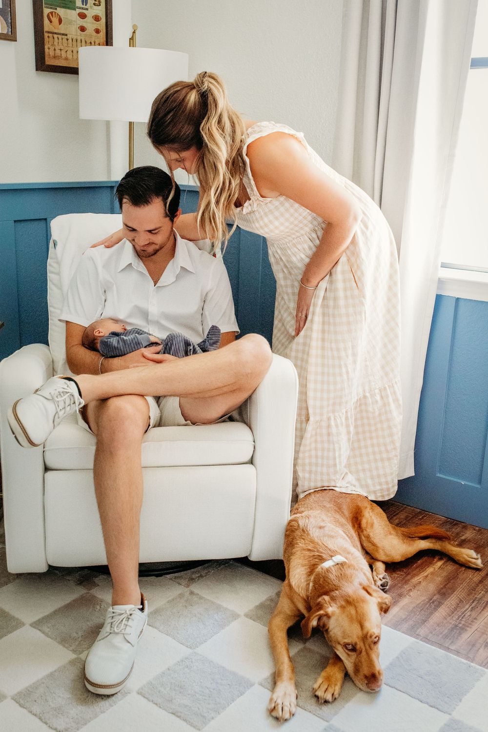 A couple sits on a white couch with their golden retriever dog in a bright room with blue walls.