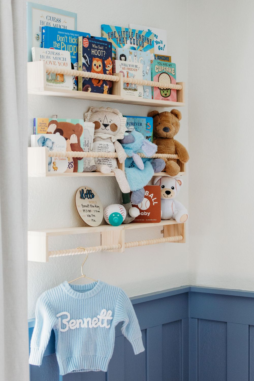 White nursery wall decorated with floating bookshelves displaying children's books, stuffed animals, and a blue sweater below in Watauga