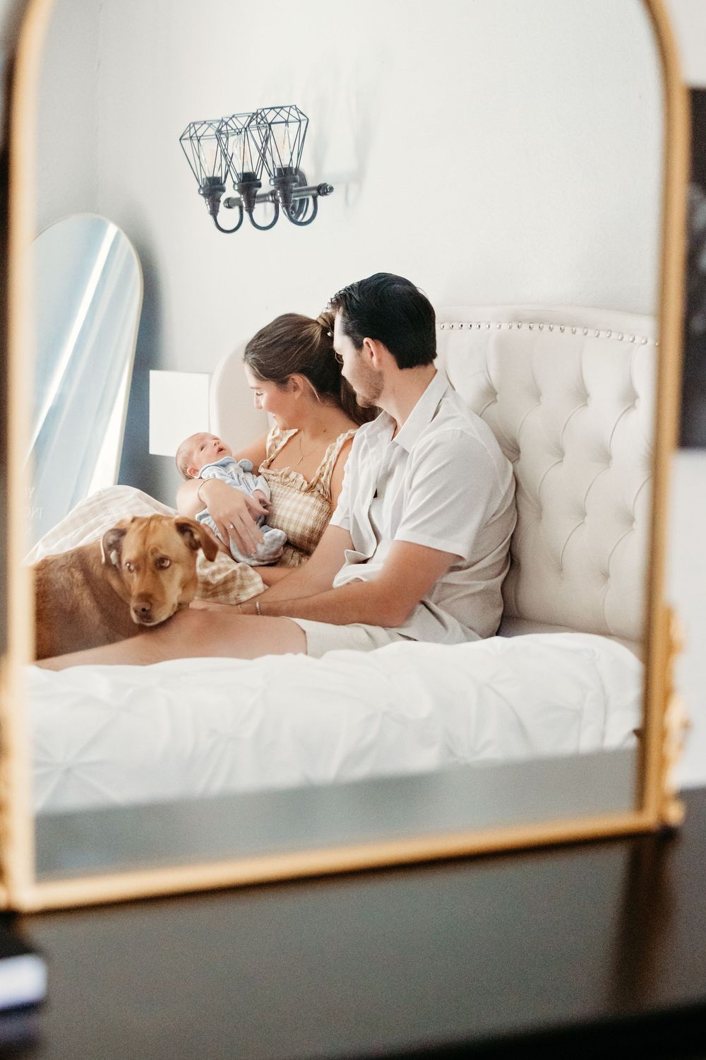 A couple and their dog relax together on a white tufted bed while reflected in multiple mirror panels.