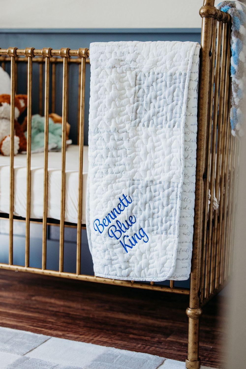White quilted baby blanket with 'Bennett Blue King' embroidered text hanging on a vintage brass crib rail.