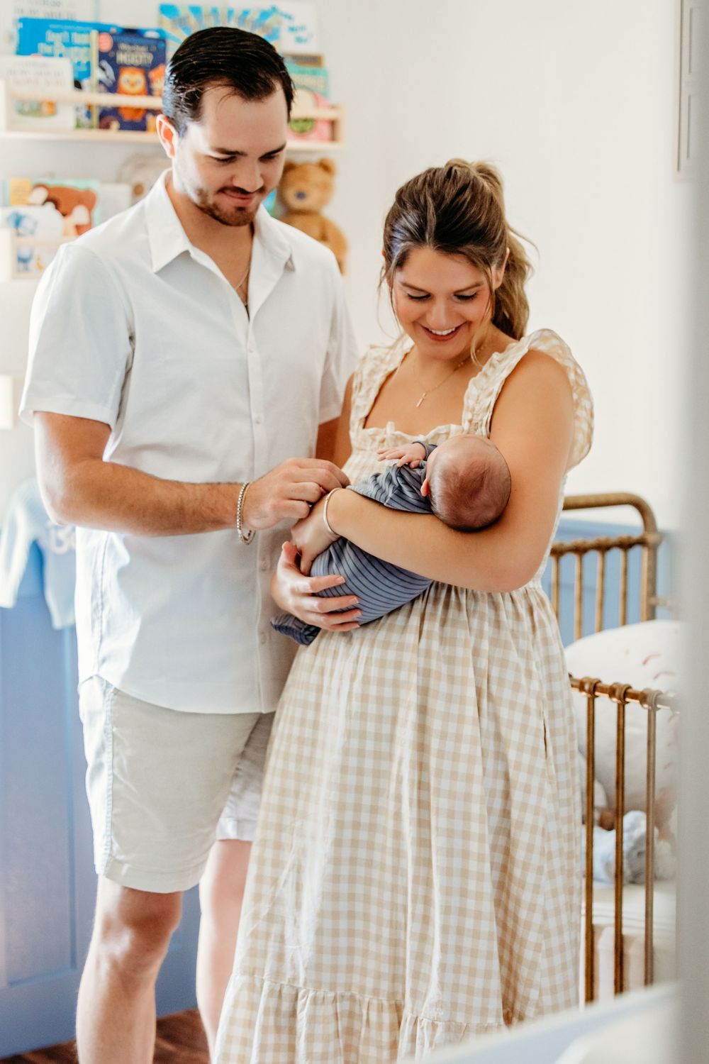 A couple in white clothing shares tender moments with a newborn baby in a sunlit nursery.