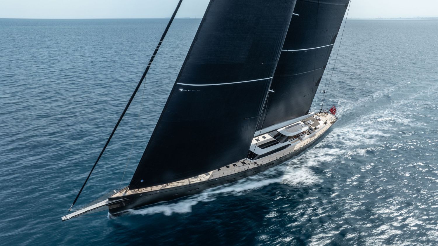 Racing sailboat cutting through ocean waves with black sails against blue horizon.