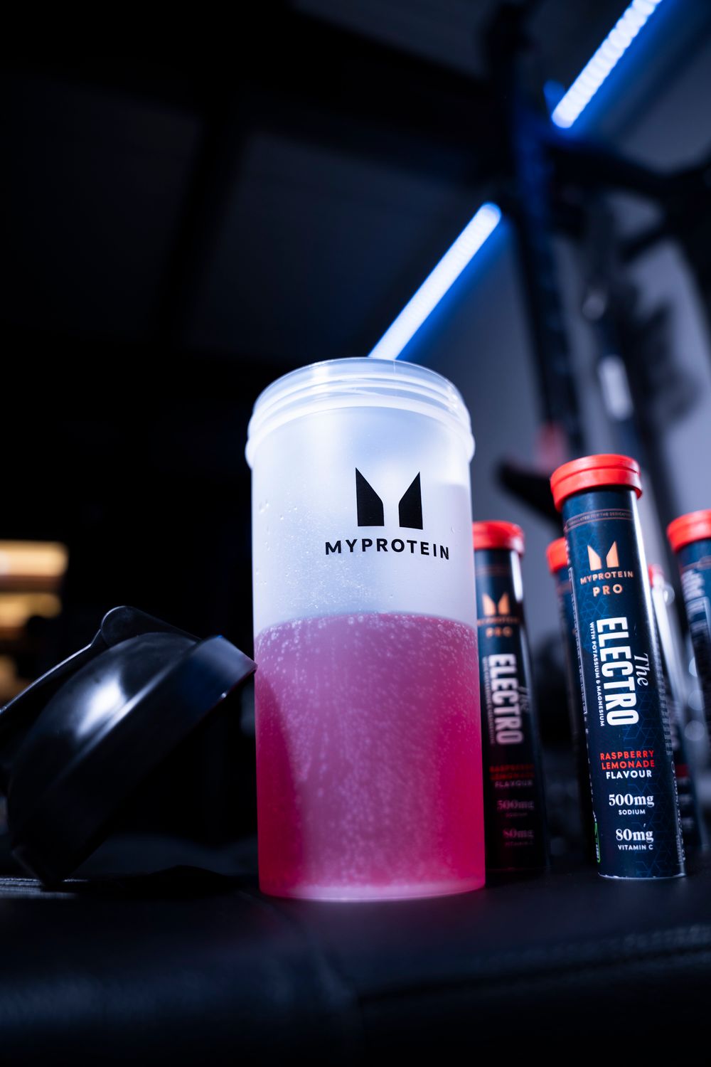 Close-up of a shaker bottle with pink liquid in a dark, neon-lit gym; commercial fitness product photography.