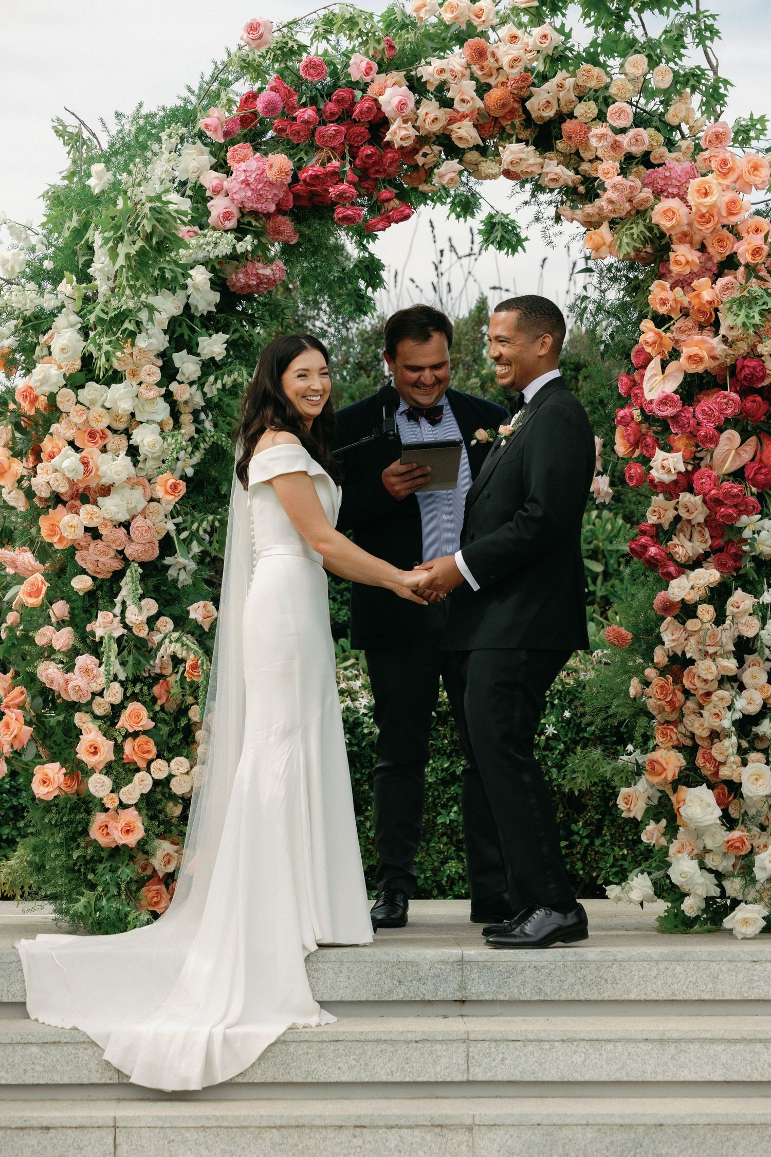 Wedding ceremony under a stunning floral arch with pink and peach roses and greenery during an outdoor celebration.