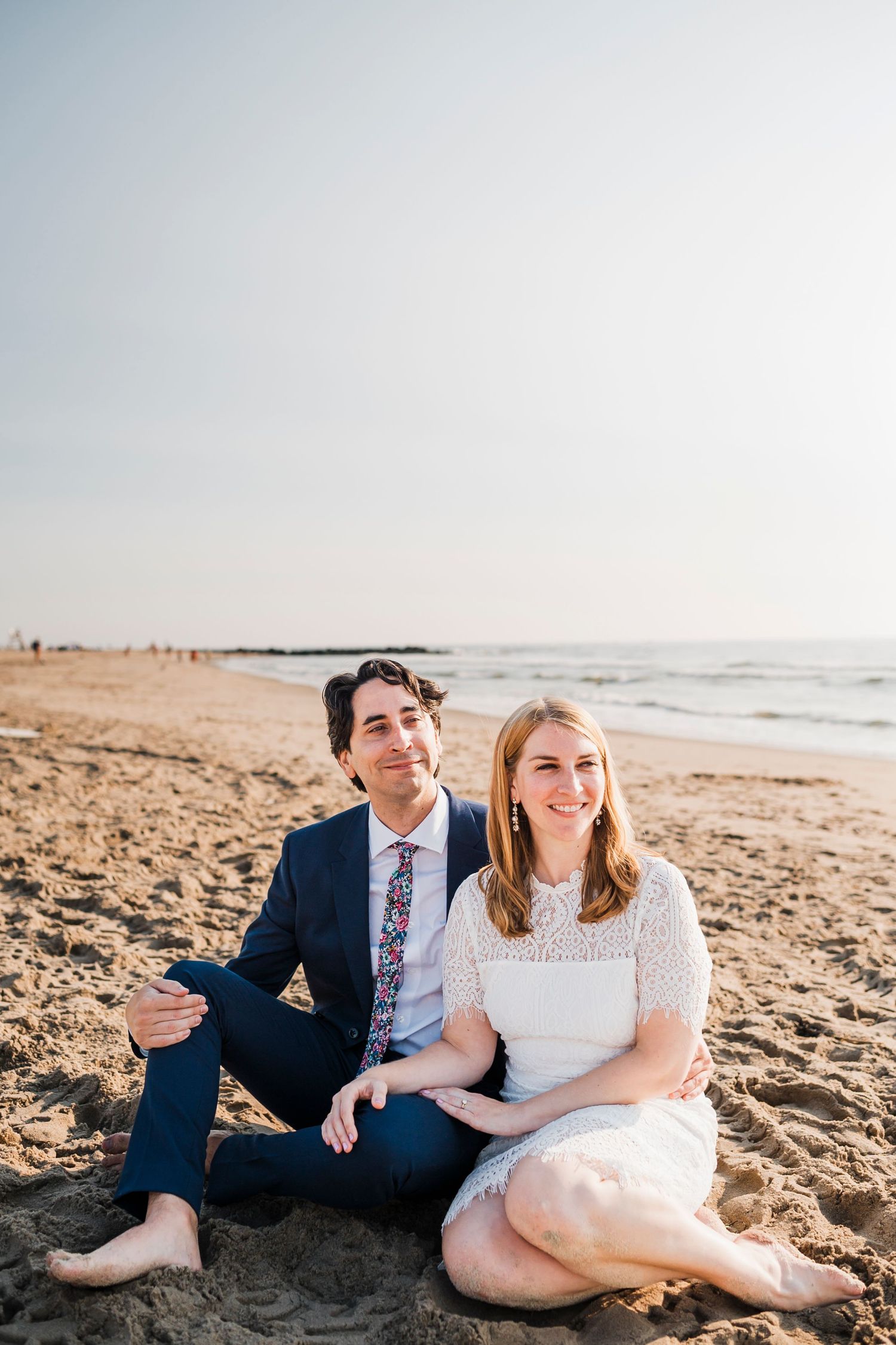 A couple sits together on a sunny beach wearing formal attire and casual bare feet.