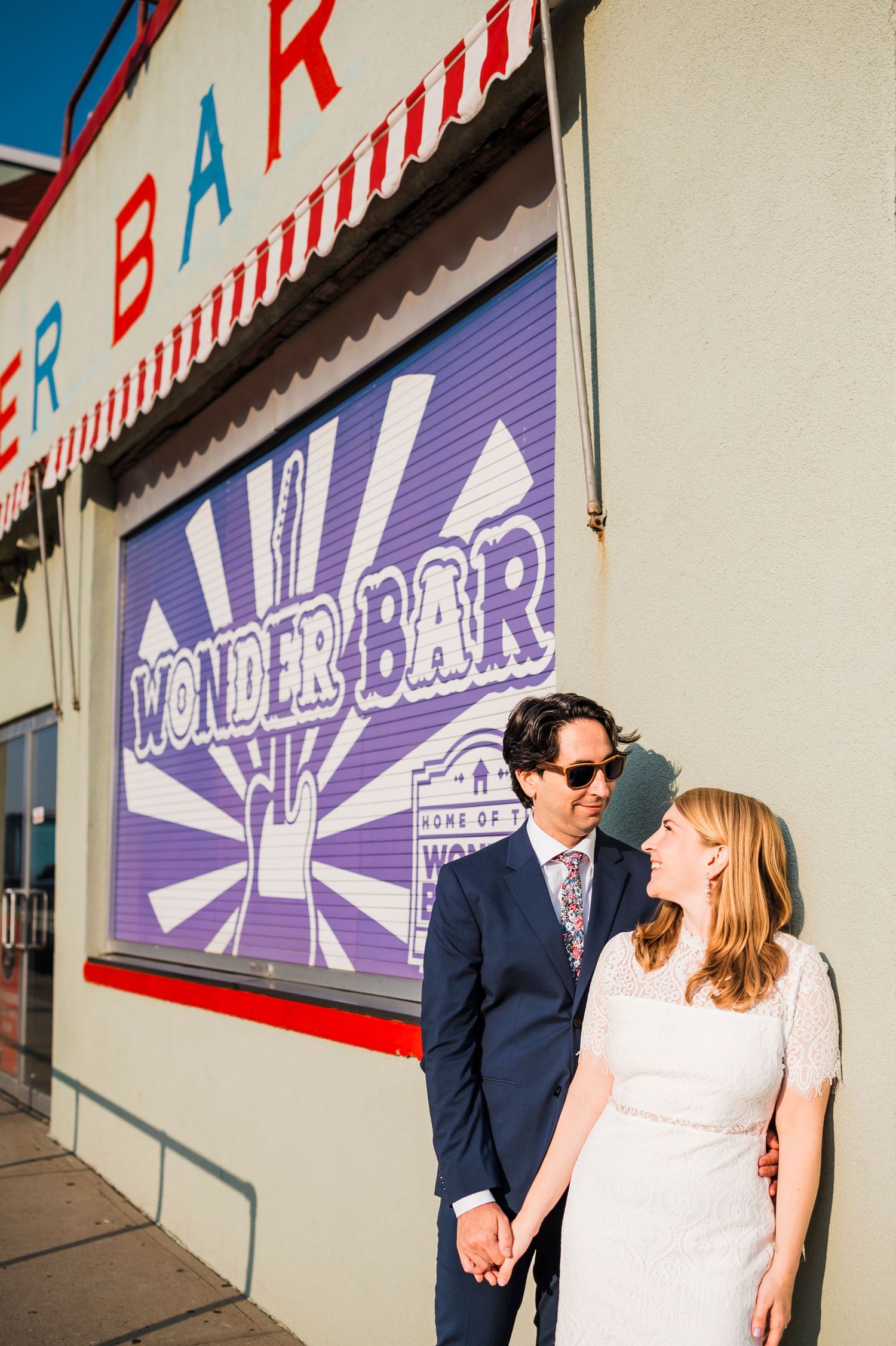 A couple poses together against a retro-style storefront with purple signage and striped awning.
