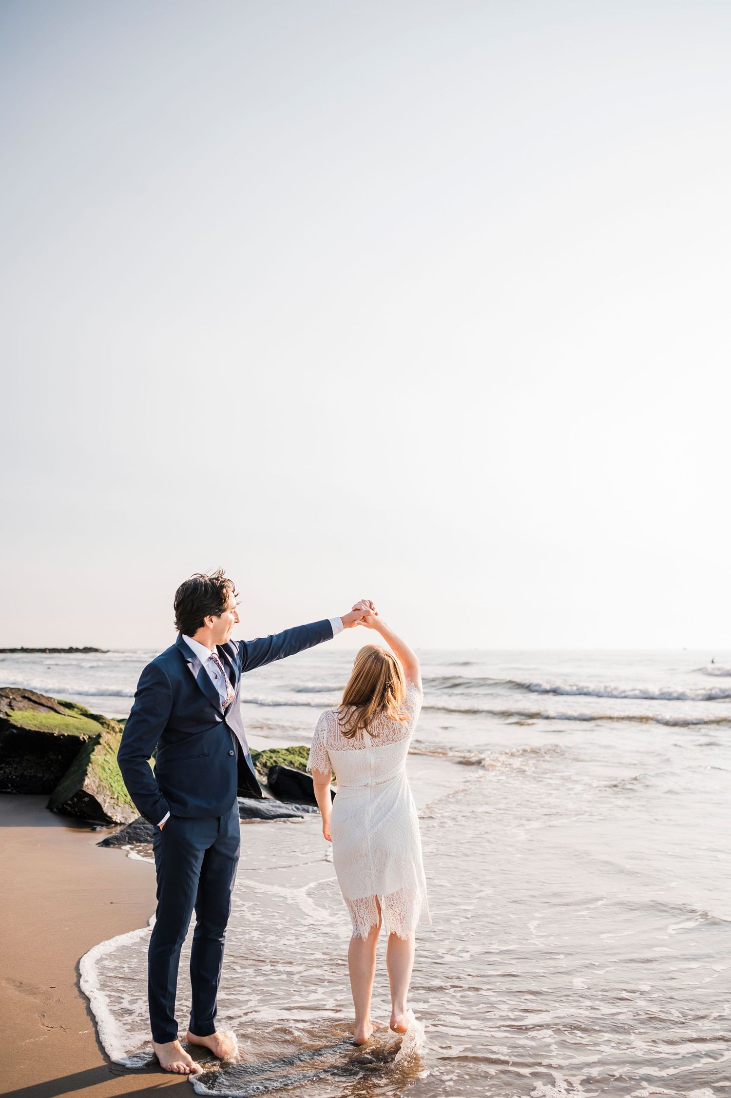 A couple walks along the beach shoreline at sunset holding hands.