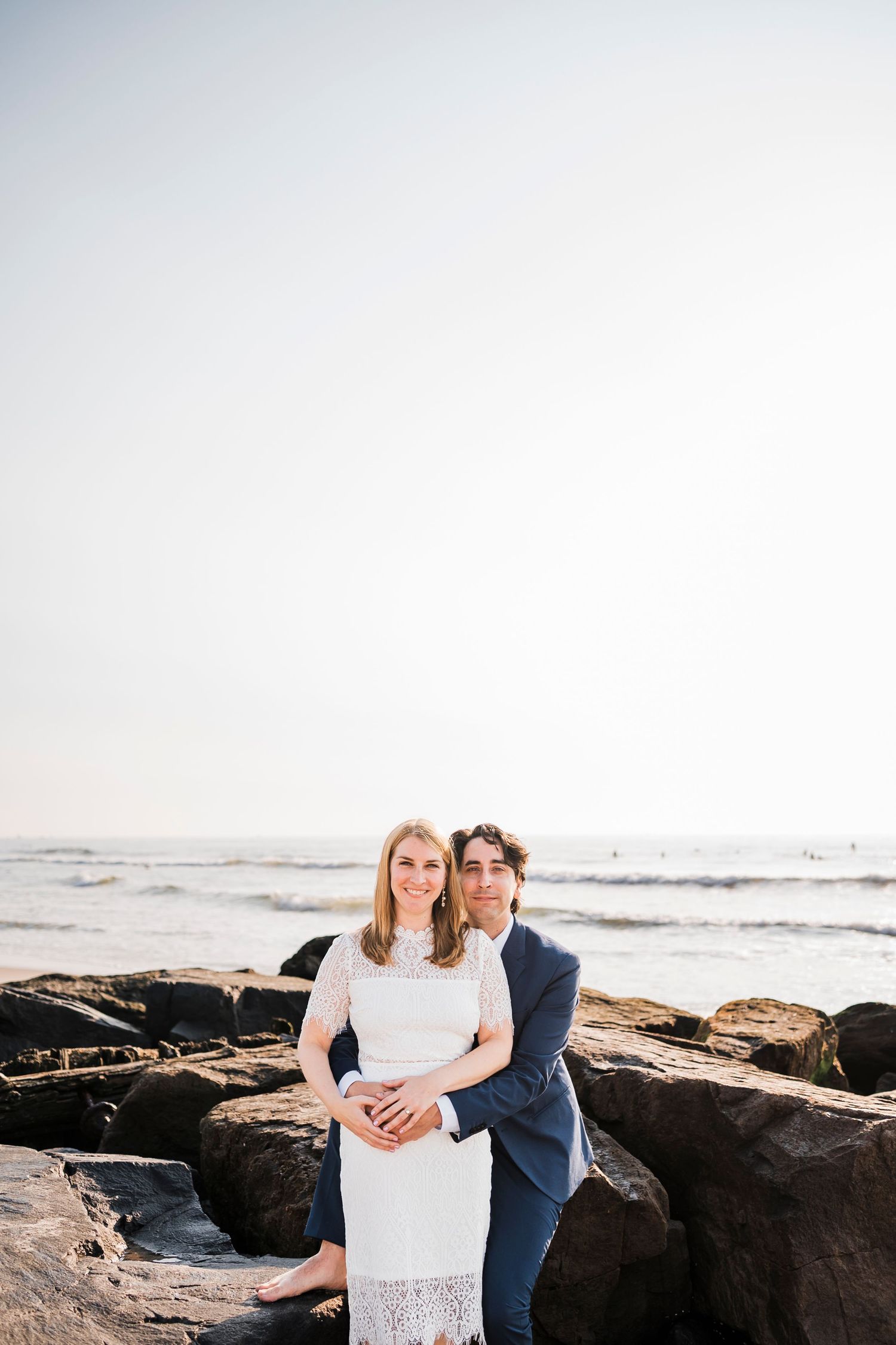 A romantic couple sits together on coastal rocks by the ocean.