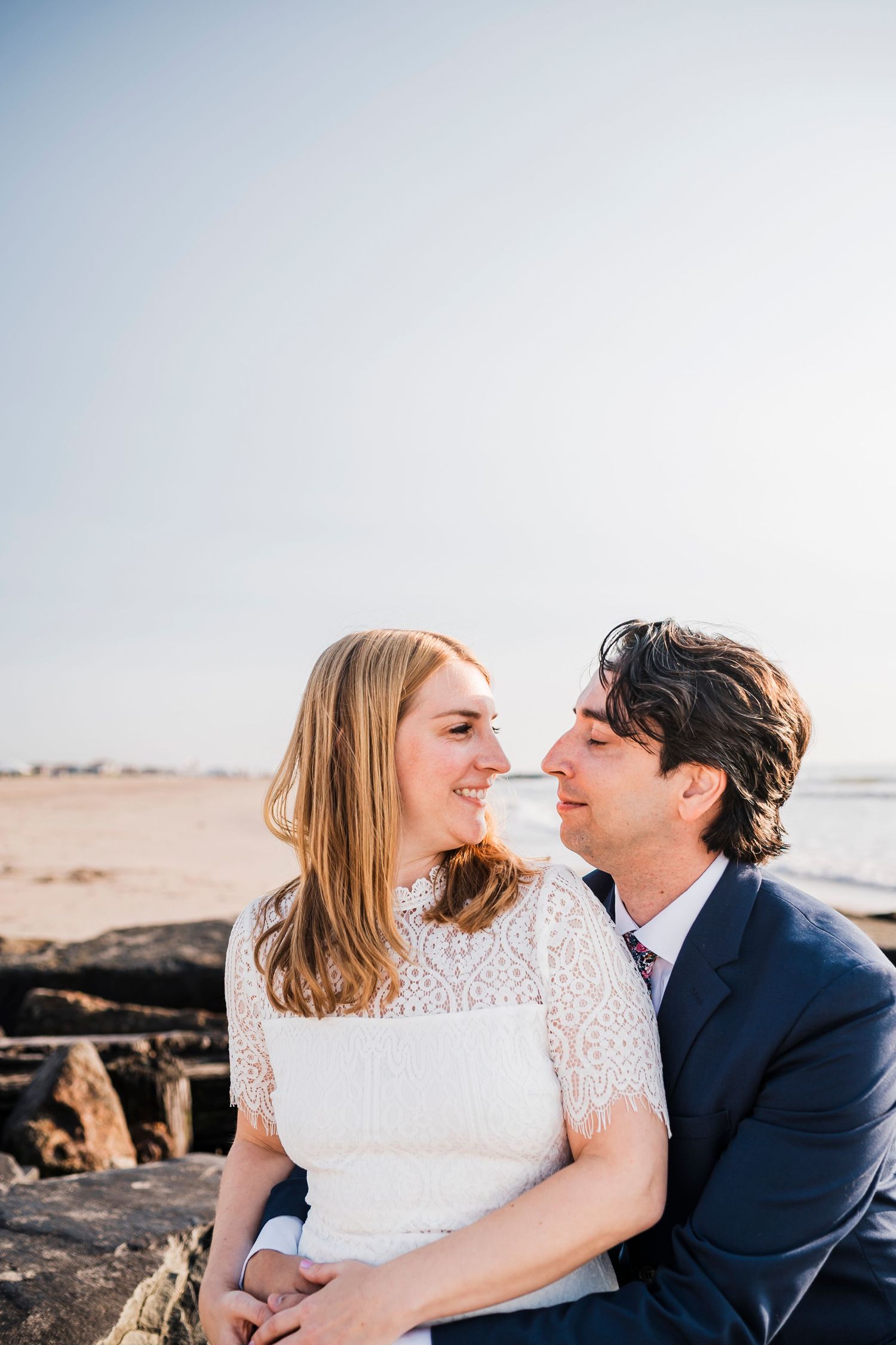 A romantic couple sits on coastal rocks, sharing an intimate moment in their wedding attire.