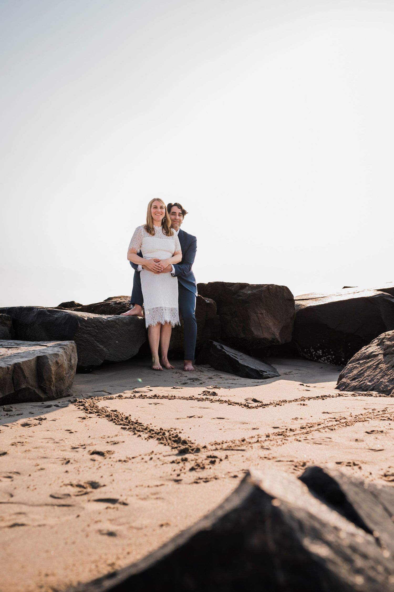 A couple stands together on a rocky beach with dark boulders and sand beneath overcast skies.