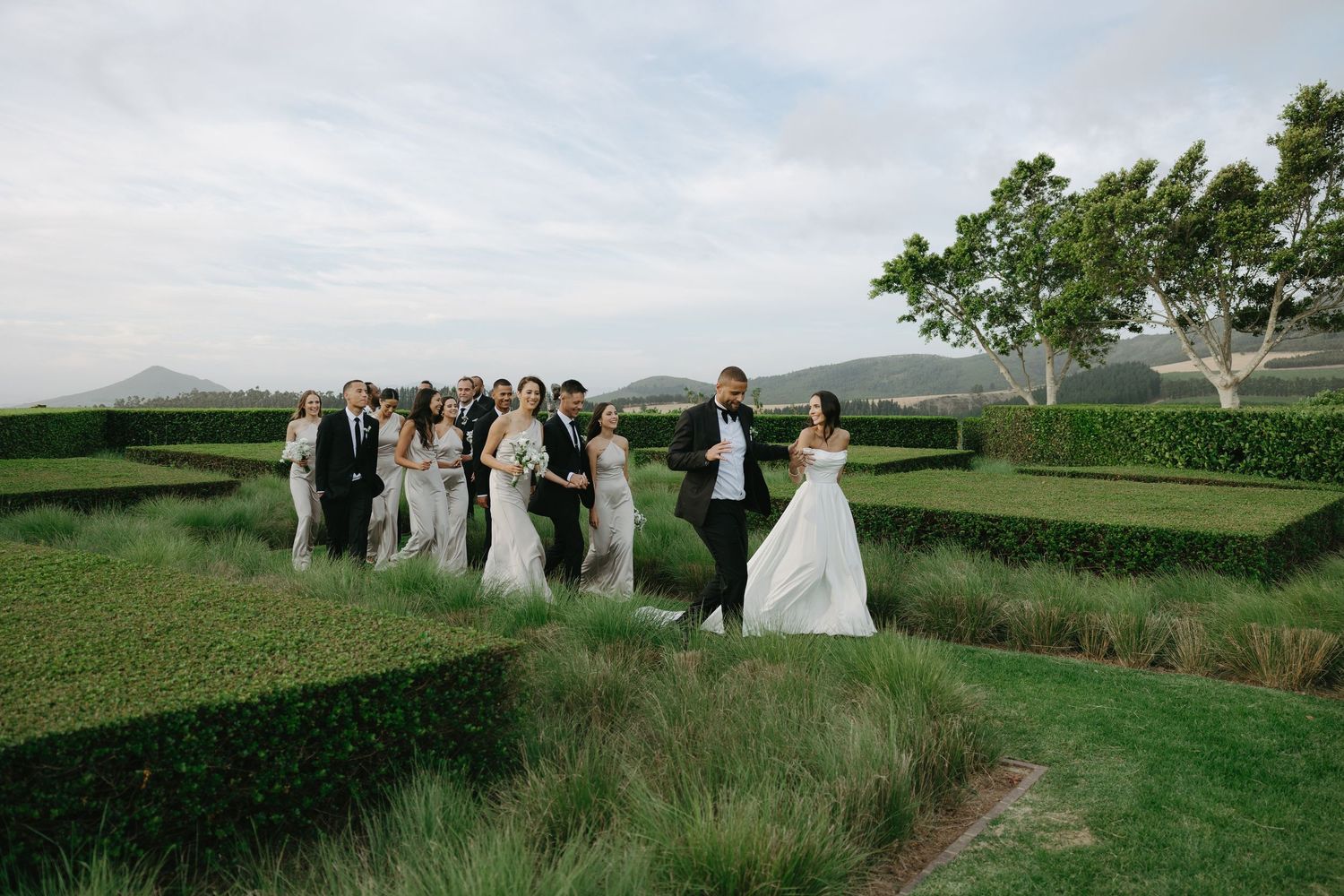 A wedding party walks through a lush green garden maze with hedges under a cloudy sky.