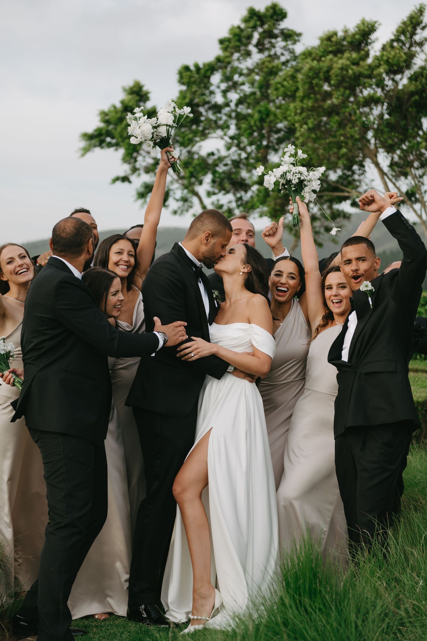 Wedding party celebrates joyfully in a row outdoors with bridesmaids in beige and groomsmen in black suits.