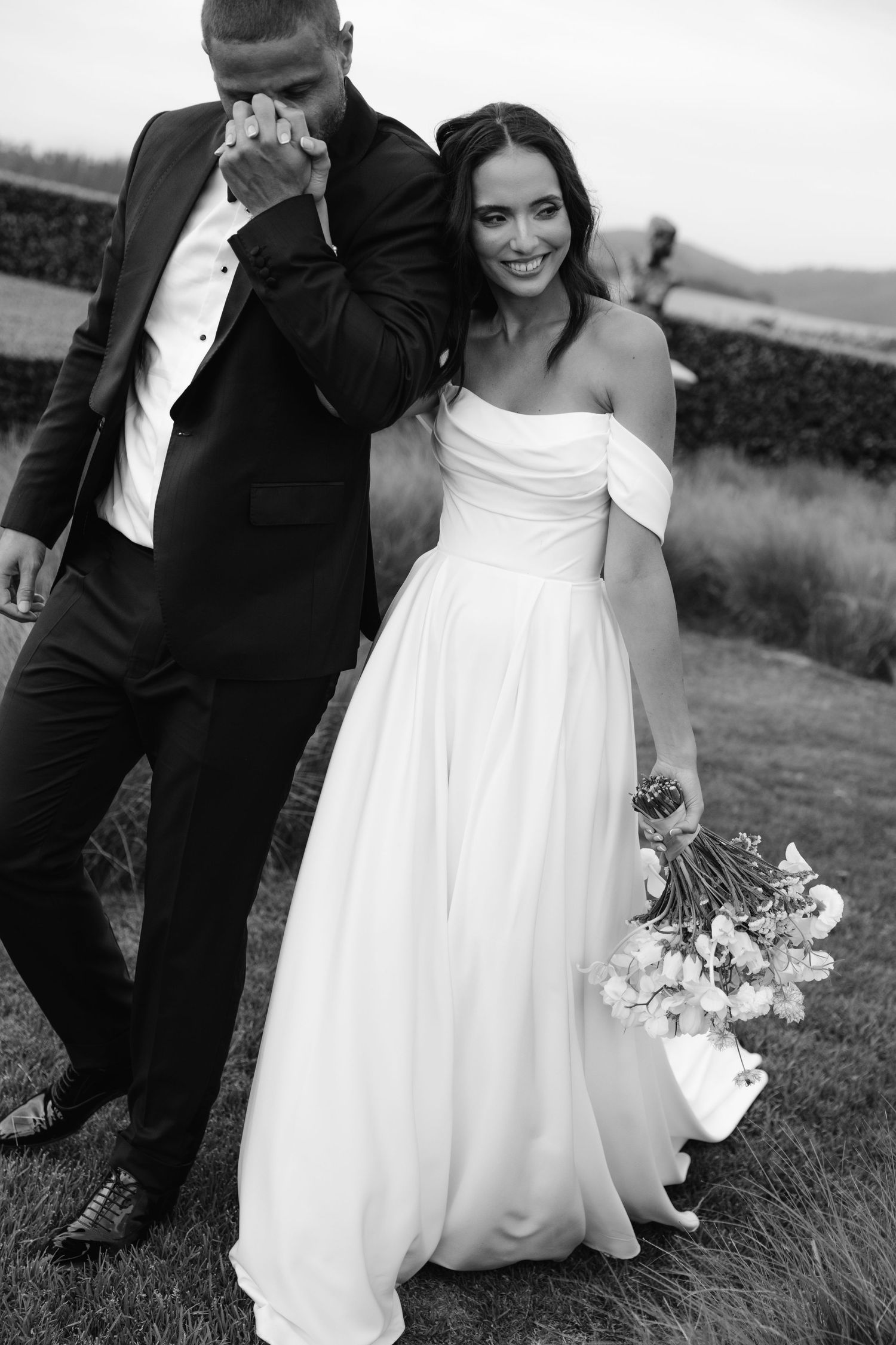A black and white wedding photo showing a couple outdoors, with the bride in an elegant off-shoulder gown holding a bouquet.