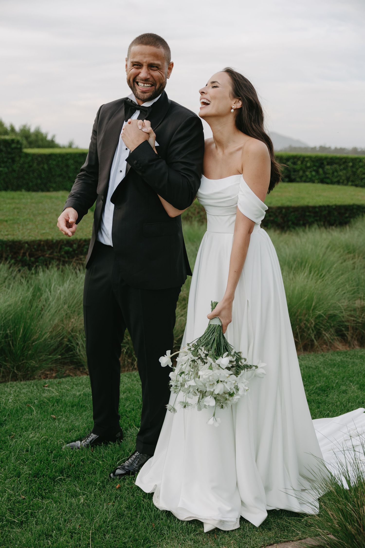 A joyful wedding couple shares candid laughing moments together in a lush green outdoor setting.