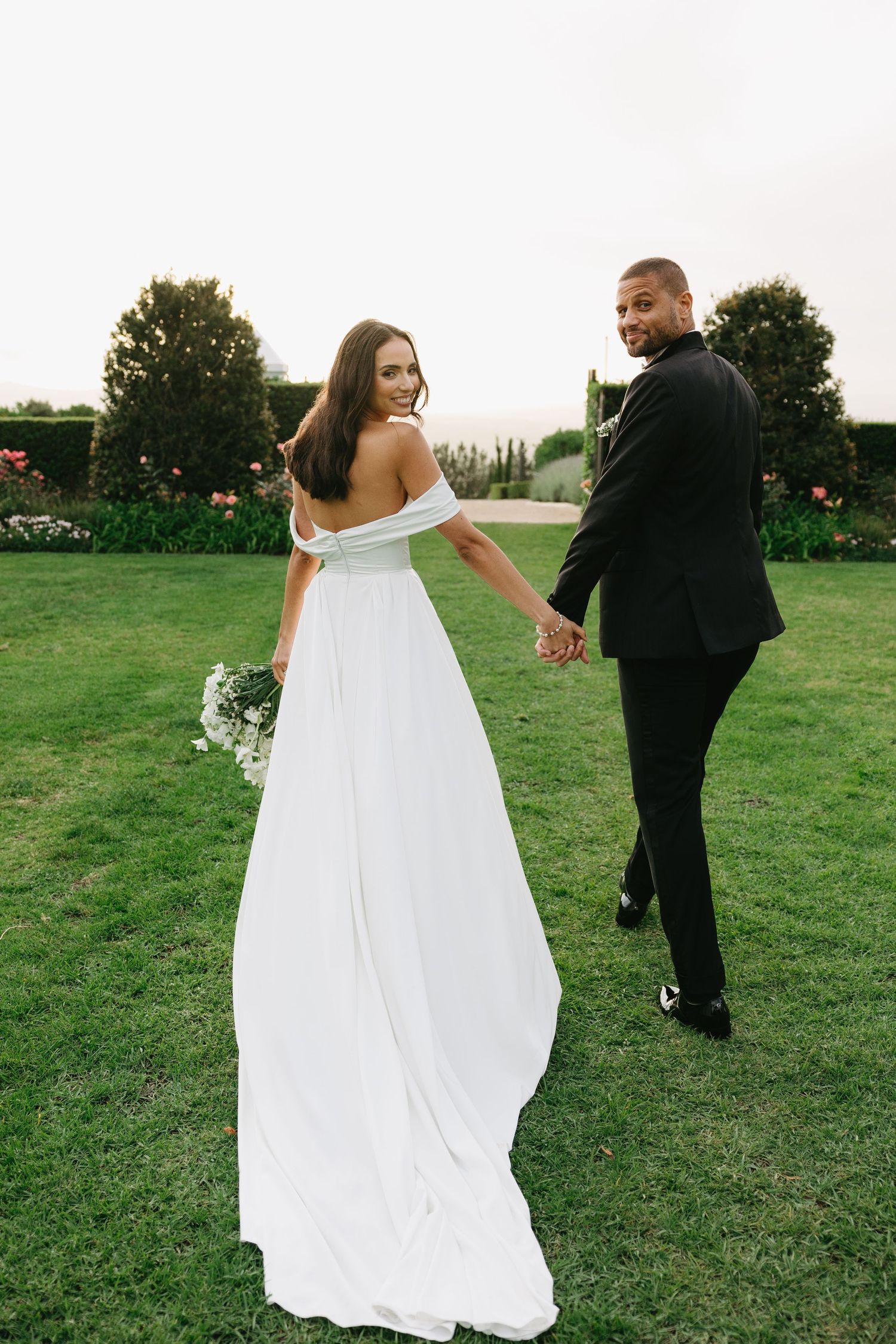 A couple in wedding attire walks together on a grassy field during a photo session.