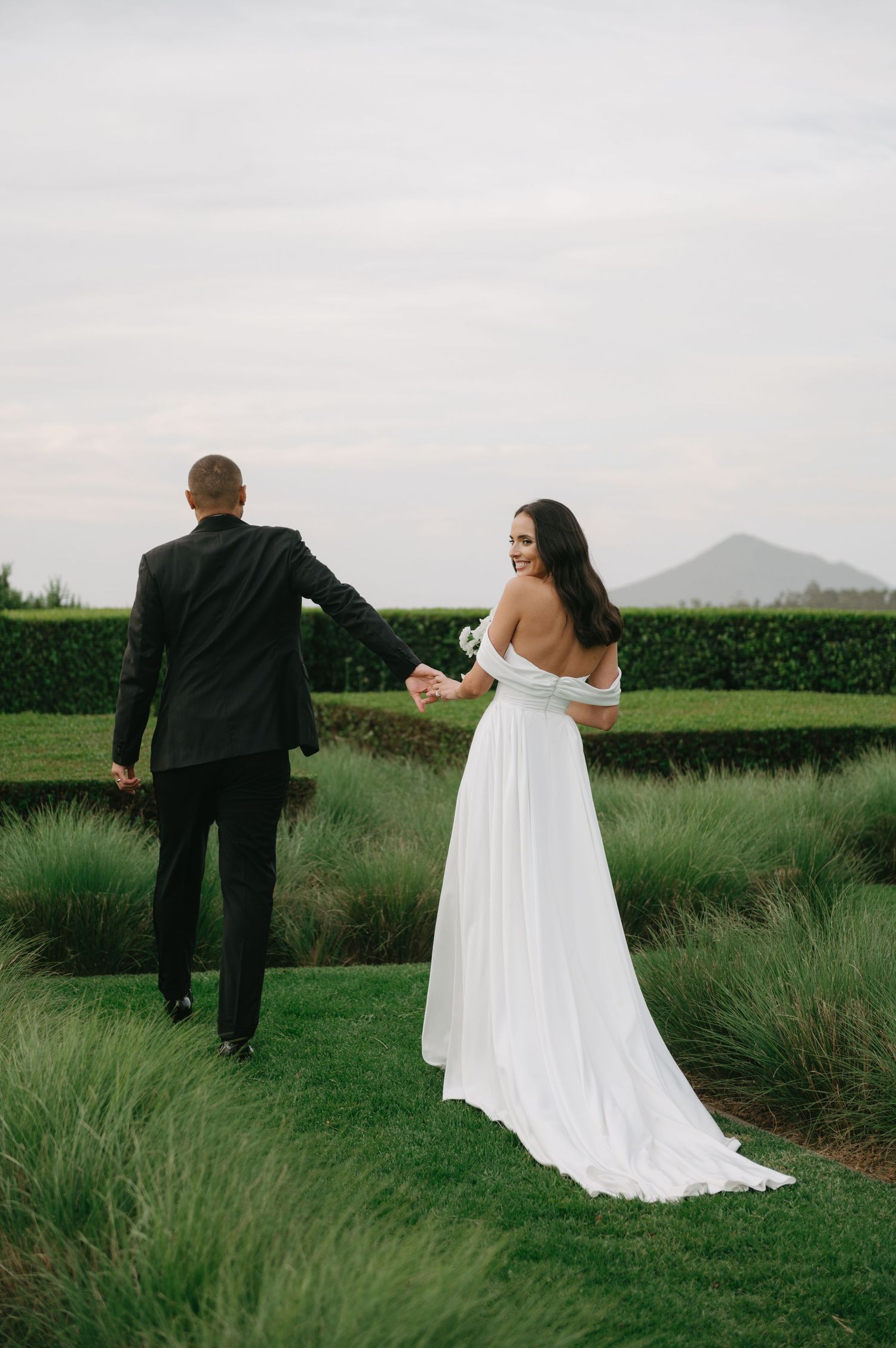 A couple in wedding attire walks through a lush green field with mountains in the background.