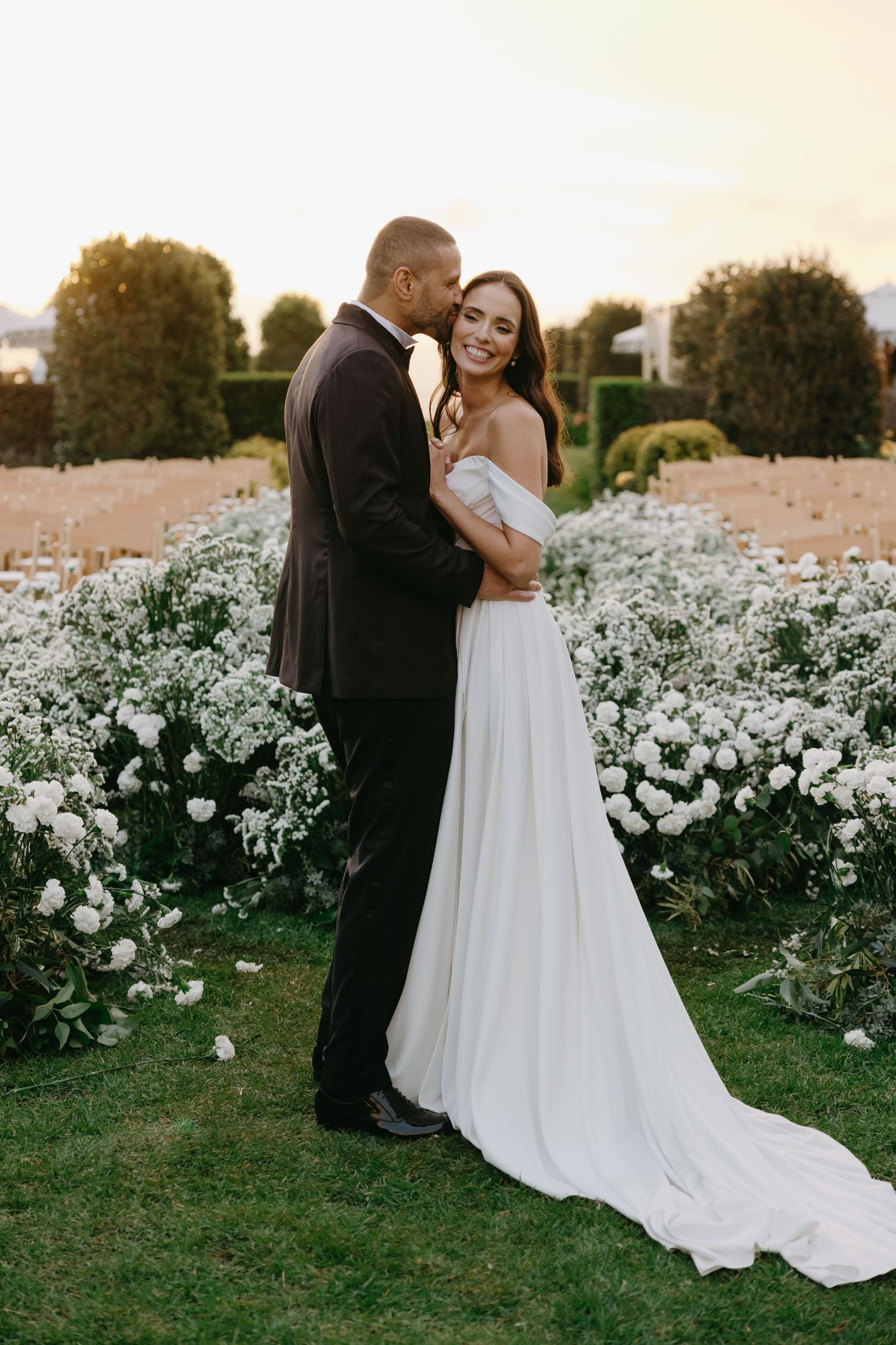 A couple embraces in a romantic sunset moment surrounded by blooming white flowers in a garden setting.