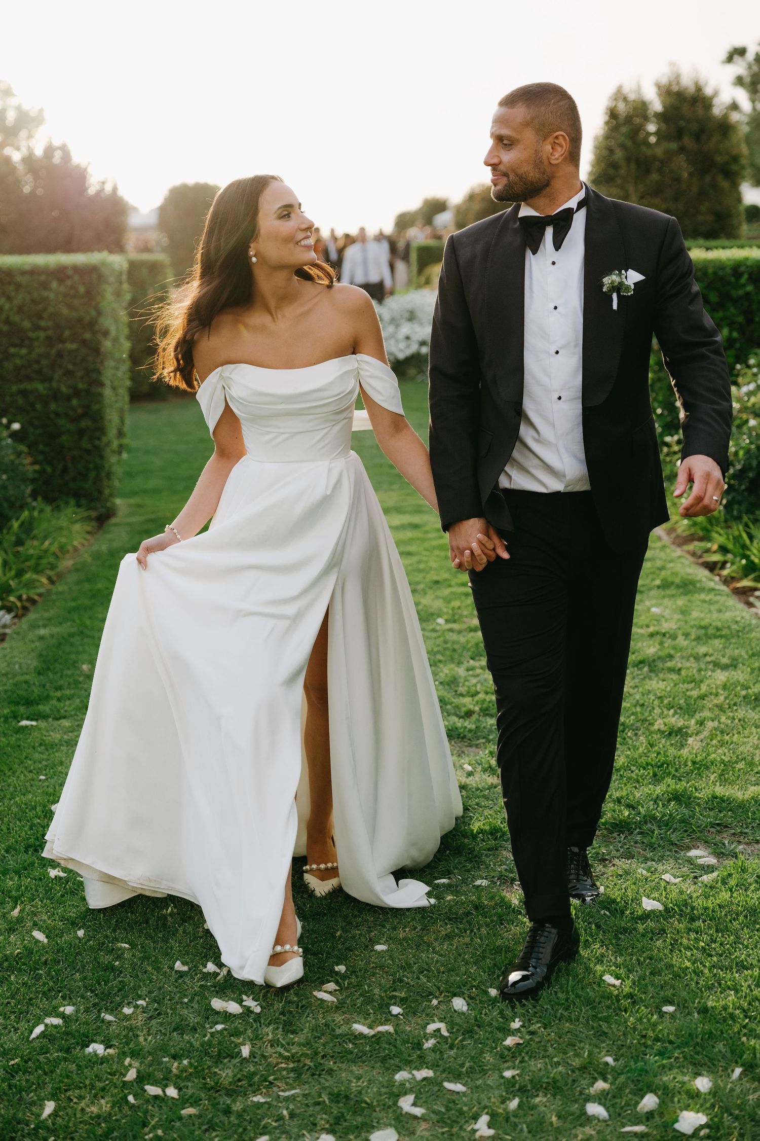 A couple in formal wedding attire walks playfully through a grassy field during golden hour.