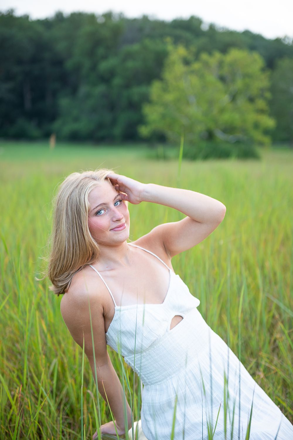 A woman in a white dress reclines thoughtfully in a lush green field of tall grass on a summer day.