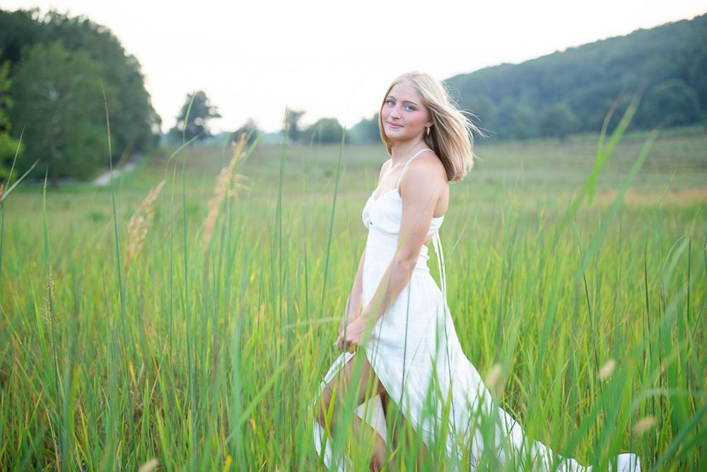 Woman in flowing white dress walks through tall grass in a summer meadow with mountains in background.