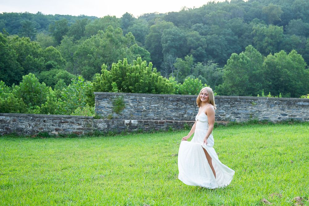 A person in a white dress twirls gracefully across a lush green meadow with stone walls and trees in the background.