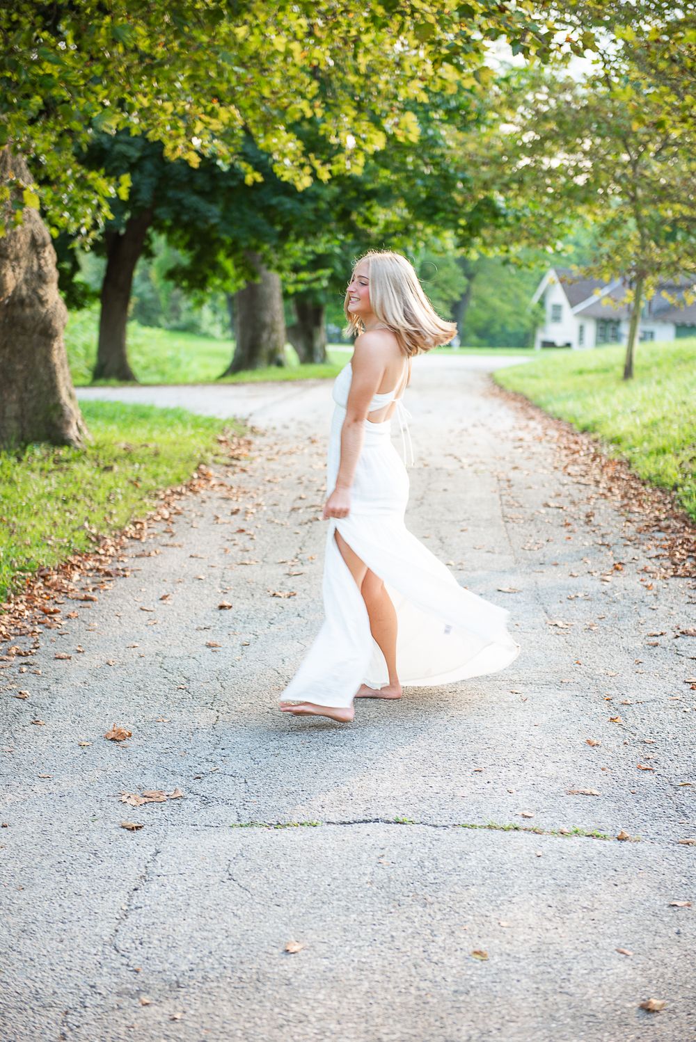 A figure in a flowing white dress twirls gracefully on a tree-lined gravel path during a sunny autumn day.