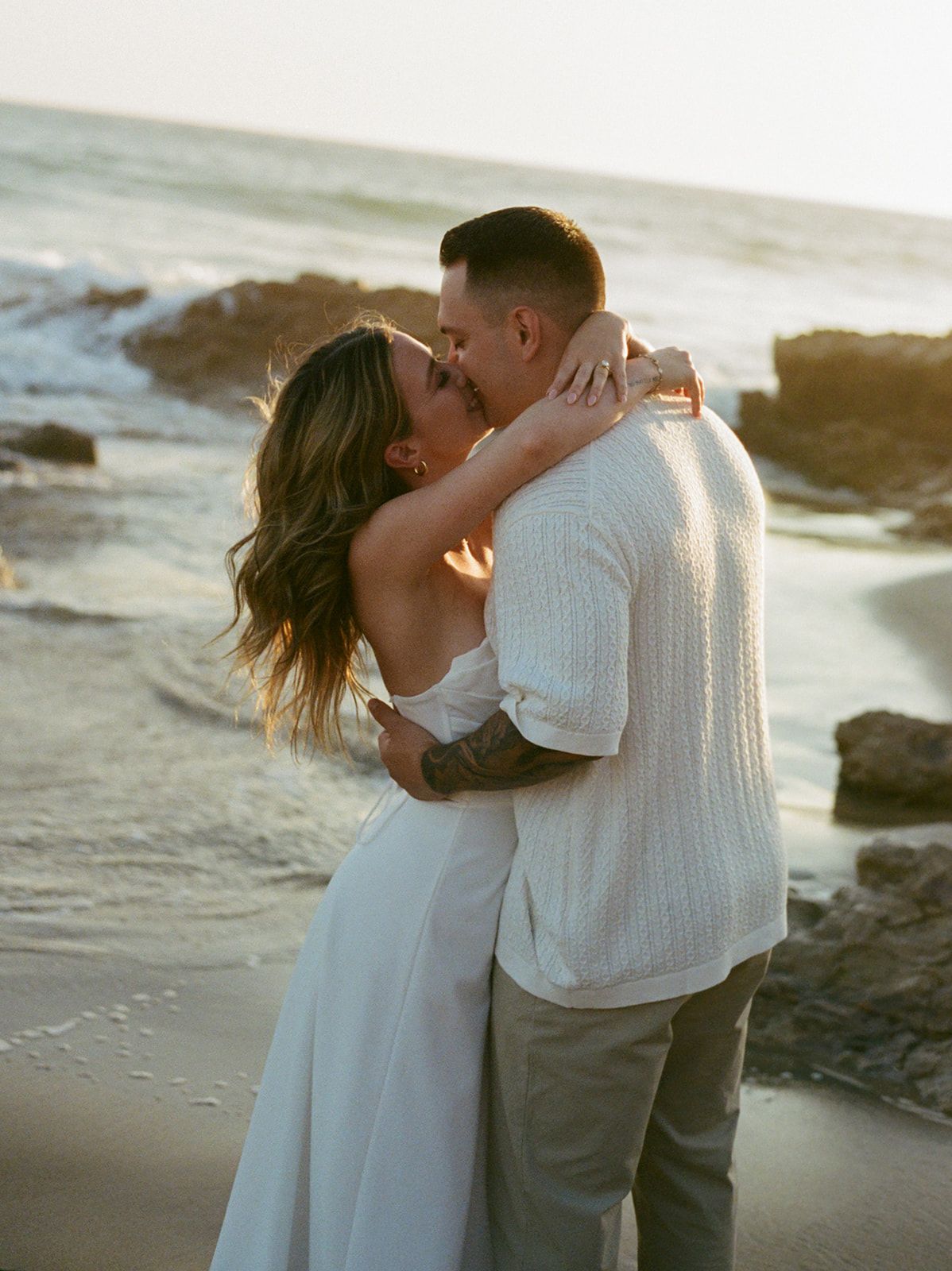 A couple stands together on rocky coastal cliffs at sunset overlooking the ocean waves and rugged shoreline.