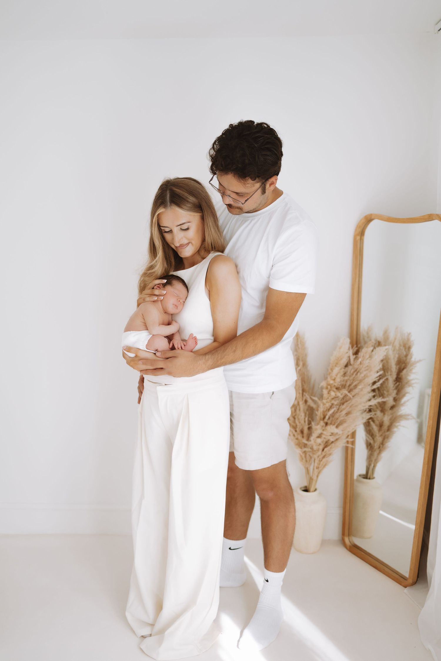 A couple in white outfits tenderly holds their newborn baby in a minimalist room with pampas grass decor.