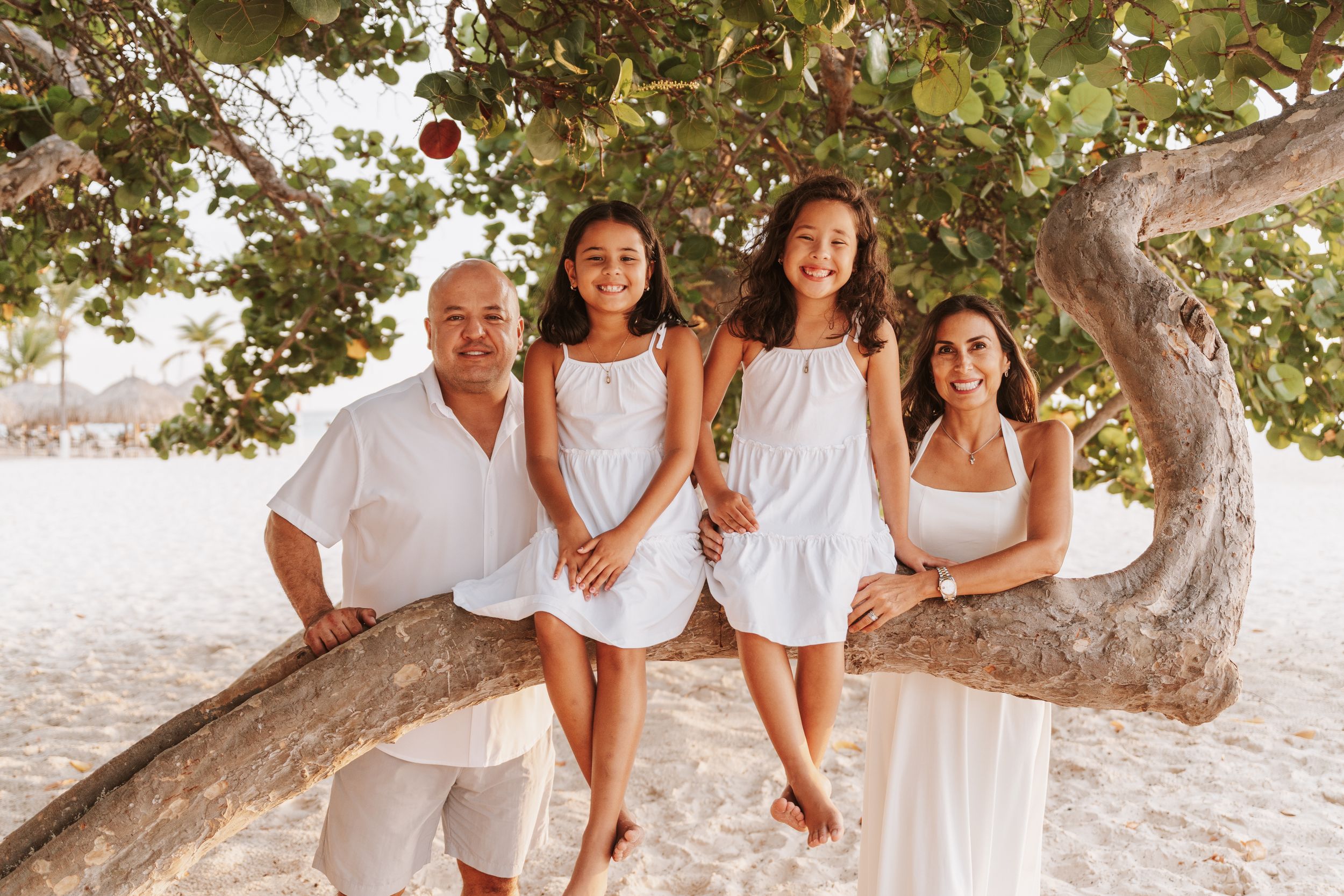 A family wearing white outfits poses together on a tropical beach near a large tree during a sunset photoshoot.