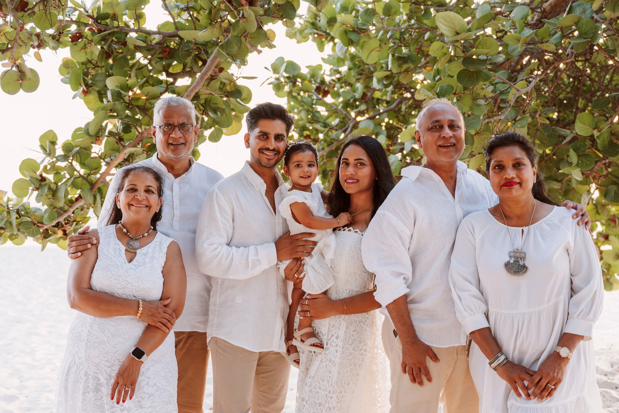 A large family group wearing white outfits poses together outdoors under green foliage for a casual photo session.