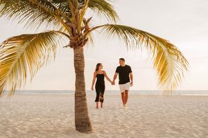 A couple walks along a tropical beach with palm trees during sunset creating silhouettes against the golden sky.