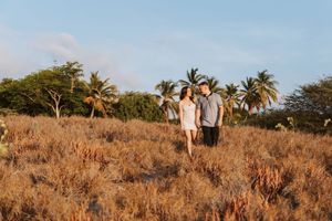 A couple poses together in a golden field with palm trees and ocean views in the background.