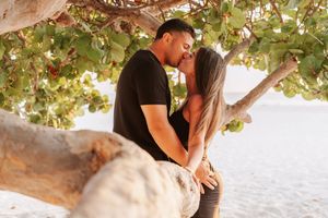 A couple shares a romantic moment under a tree by the ocean during sunset.