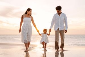 A couple walks barefoot along a sandy beach at sunset holding hands in white summer attire.