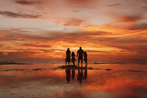 Silhouettes of people standing together on a beach at sunset with vibrant orange and red sky reflecting on wet sand.